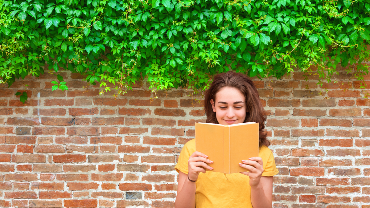 Woman reading book in front of brick wall.